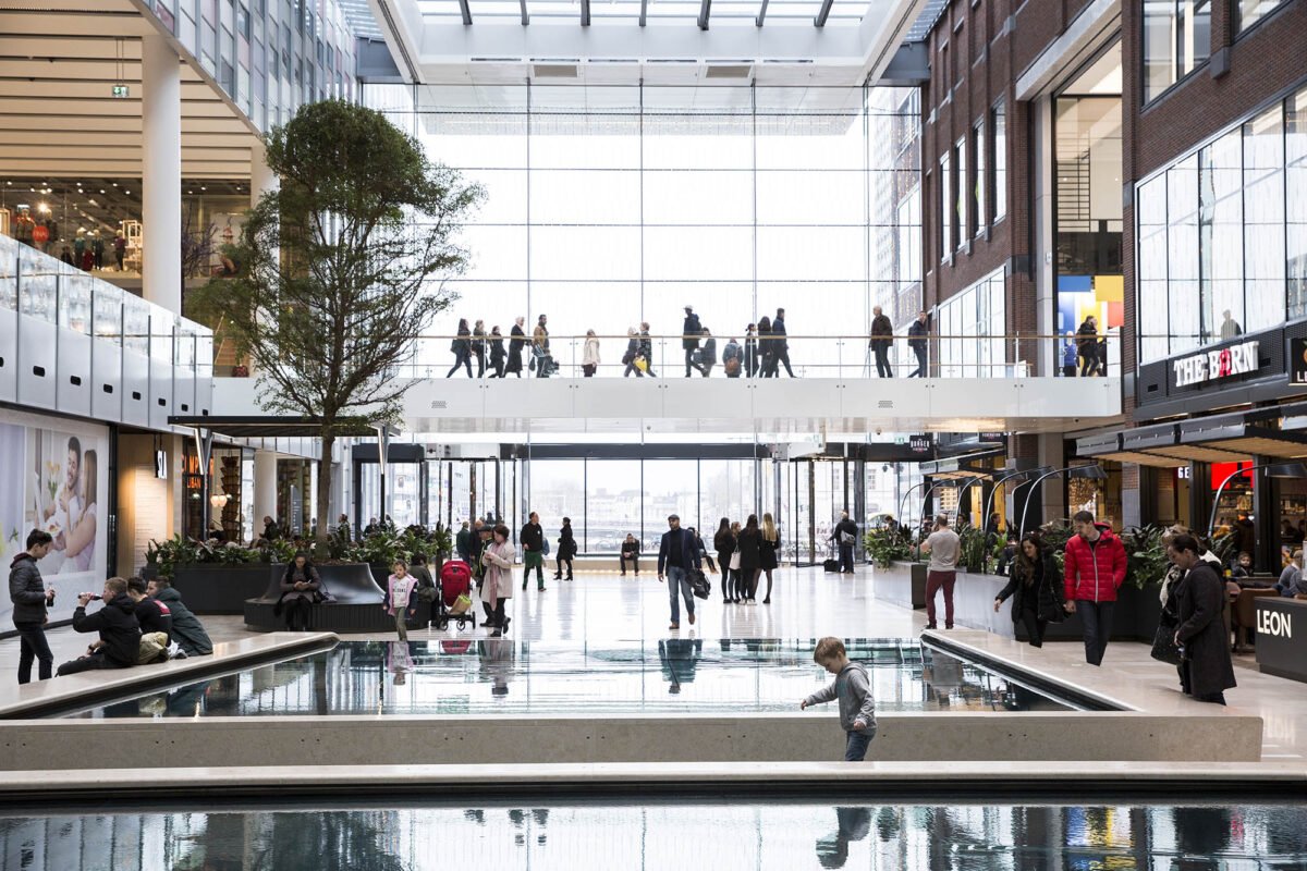 A bright indoor public room at Hoog Catharijne in Utrecht: a glass roof, a footbridge, a shallow pool, and people walking between shops.
