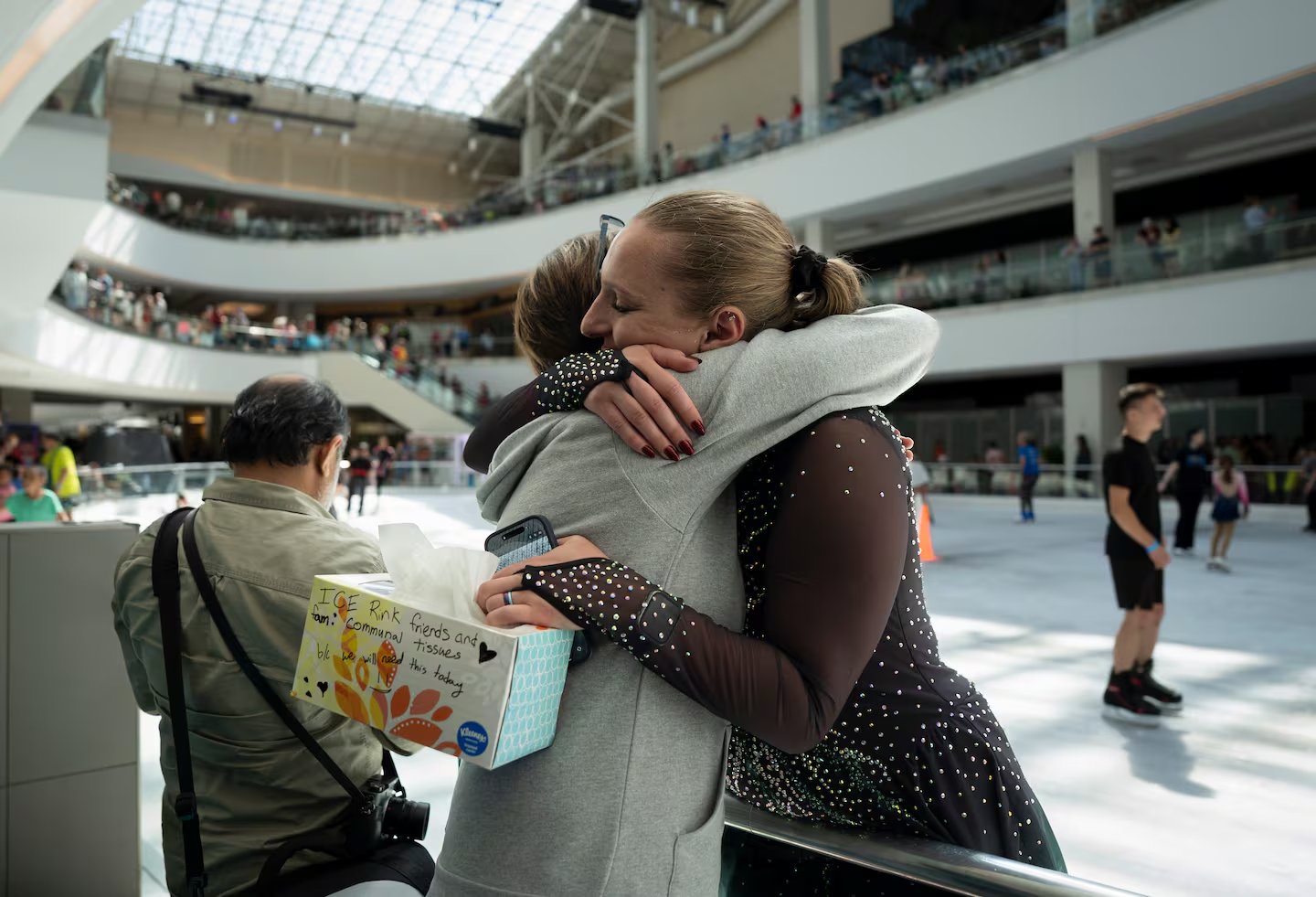 Two people embrace at the rink rail, one in a skating dress, while other skaters and crowded balconies fill the mall. Photograph published by OPB.